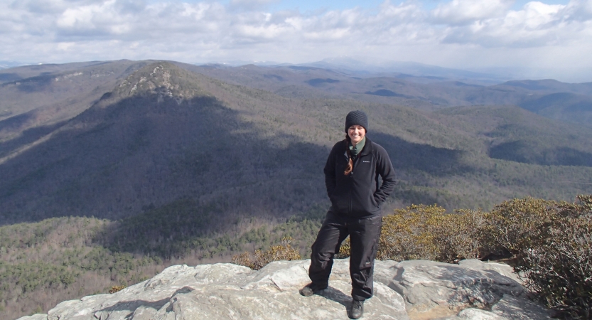 A person stands on a rock overlook and smiles at the camera. Behind them are the vast blue ridge mountains. 